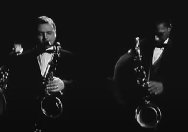 Screenshot from 1960 video of Stan Getz and John Coltrane on stage in Germany, both in bow ties and formal suits holding their tenor saxophones. At this moment in the video, Getz is playing and Coltrane is looking down.