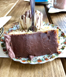 A triangular piece of dark brown cake with brown icing with a hemispherical scoop of white ice cream with brown chocolate shards on a blue plate on a rectangular light brown table on a bright background 