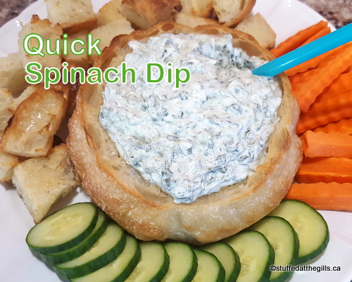 Quick Spinach Dip in bread bowl surrounded by toasted bread pieces, carrot sticks and cucumber rounds.