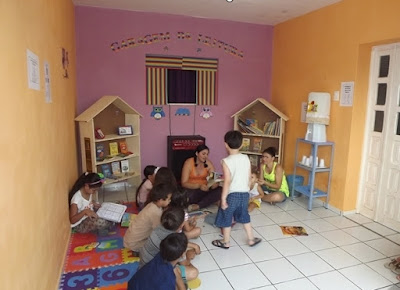 Estantes  de madeira em formato de casa com livros pratileiras.  A frente mulher lendo historias para crianças. 