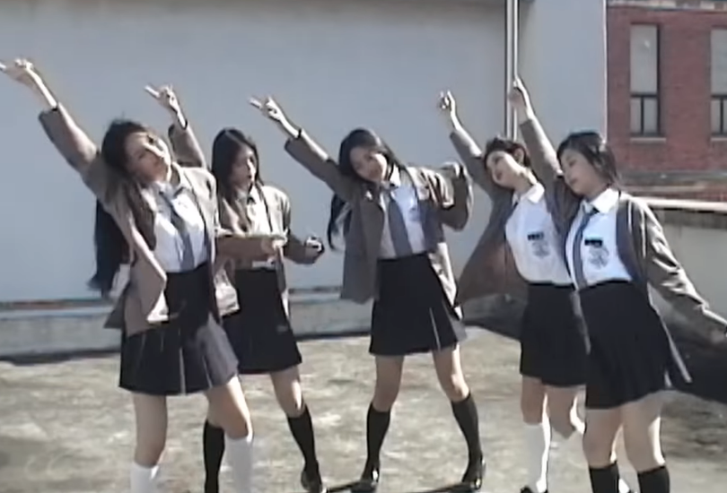 Camcorder footage of the members of NewJeans dancing on a rooftop, wearing their high school uniforms.