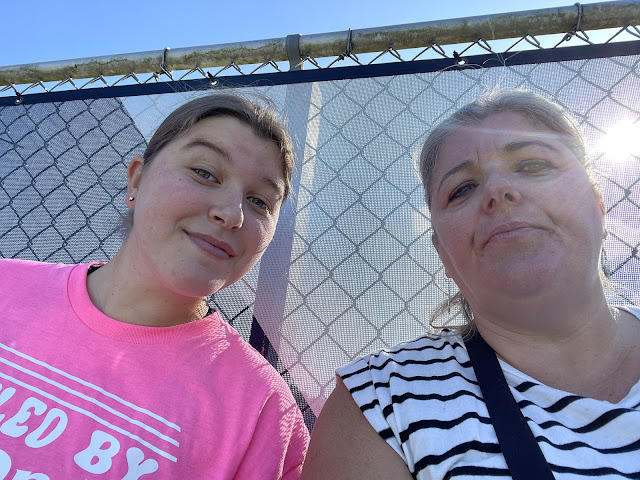 A selfie of Elizabeth and myself sitting up against a chain link fence on the top row of some bleachers. Elizabeth is wearing her pink 'fueled by dr. pepper' tshift and I am wearing a white tshirt with small black stripes running horizonally across my body. We are leaning into each other smiling at the camera.