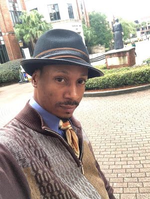 A black man with medium brown skin is slightly off center. He is wearing a half-zip sweatshirt in a brown pattern over a blue shirt with a gold and brown striped tie. He has on a gray hat with brown band. He has a black mustache and goatee. A brick pathway can be seen behind him, as can a gray statue surrounded by hedges in the distance..