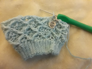 a crochet sock in progress, worked in a diamond pattern of front post stitches over a background of double crochet.  A stitch marker with a silver rose charm is clipped into the work.