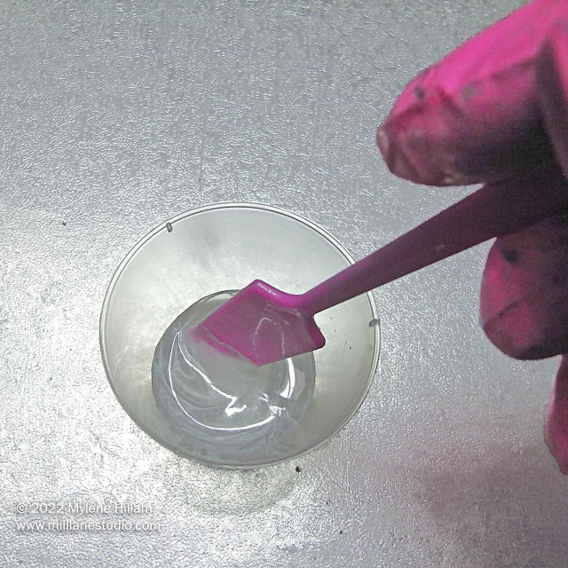 Gloved hand holding a pink plastic stirrer, stirring the white pigment into the resin
