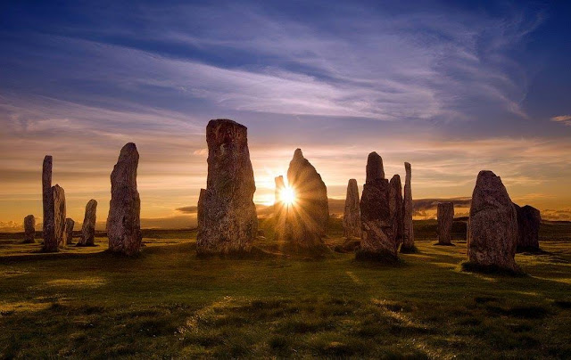 A stone circle with the setting behind it and it shining through the central stone. The ground around is green grass. It is of the Callanish Stone in Scotland, taken by Swen Stroop.