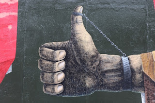 Photo of Artwork on Berlin Wall of a closed hand with a chain attached to the thumb holding up the thumb by betexion at https://pixabay.com/photos/hand-thumb-east-side-gallery-2779522/