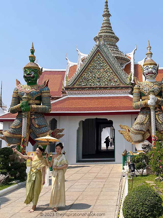 Thai women in traditional outfits walking outside Wat Arun in front of the Giant Demons, Thai women in ethnic wear with umbrella