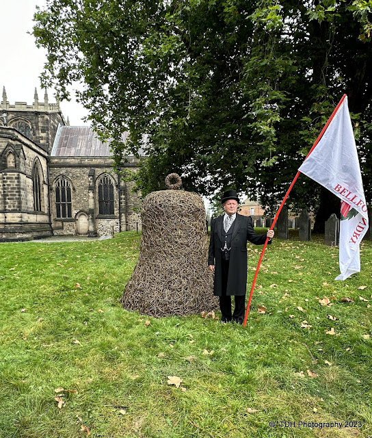 A man in Victorian gentleman's dress, with flag, in churchyard with a 7-foot willow bell sculpture