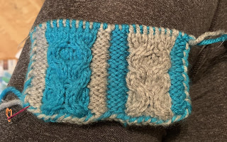 An in progress knitted test piece. Left to right with the color of the stitches: 2 gray reverse stockinette stitches, an XO cable in teal, 2 gray reverse stockinette stitches, 2 teal reverse stockinette stitches, an XO cable in gray, and 2 teal reverse stockinette stitches.