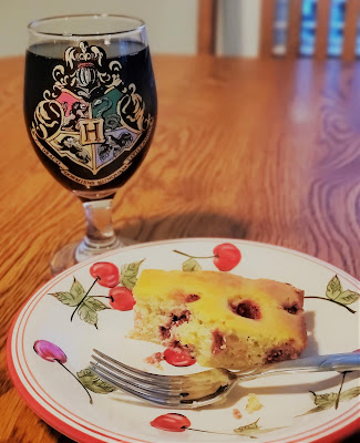 small piece of yellow cake with raspberries baked in, placed on a ceramic plate with cherries painted on it. Next to it is a wine goblet with the Hogwarts Houses painted on it. 