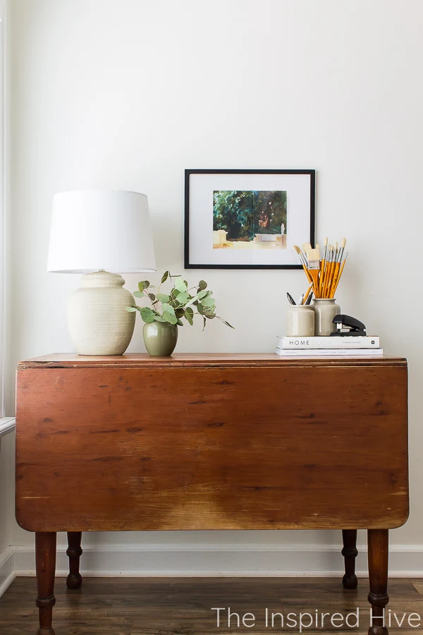 Antique drop leaf table with vintage lamp, abstract artwork, and crocks of paintbrushes