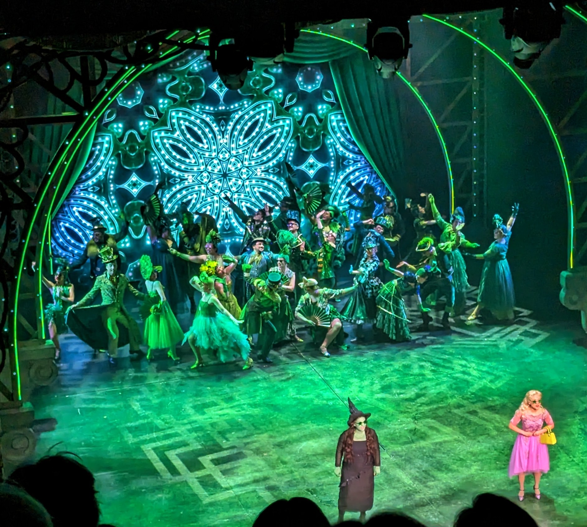 St James Theatre from the upper circle during the performance of Wicked. The lights are all set to green, the two main stars are forward and centre stage, behind them the ensemble are dressed in many different stylised Wizard Of Oz costumes.