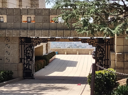 Photo from the street of a driveway leading through a gate in a Mayan-pattern concrete block house toward a terrace that overlooks Los Angeles from the hills to the north of the city. Over the gate is more Mayan pattern stone and to the left you can just make out one wall of a very large house.