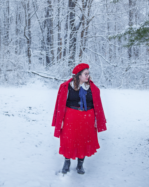 An outfit consisting of a red beret, red coat draped over the shoulders of an oversized white peter pan collar under a black sleeveless dress under a red pleated midi skirt and Doc Marten Lenore chelsea boots.