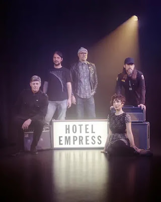 Five musicians in a dark room, standing around a lit up cinema style sign that says "Hotel Empress" on it.