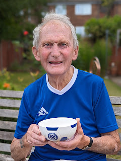 My dad a gentleman in his 80's sitting on his garden bench wearing a royal blue t-shirt with a big grin on his face and holding a blue and white cereal bowl with the Portsmouth FC emblem on the front