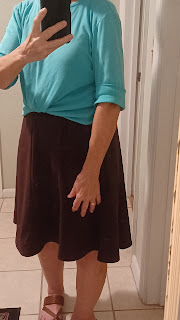 A turquoise colored pullover top is shown with the sleeves rolled up above the elbow. The front is also knotted, with the knot tucked under. It is worn with a brown A-line skirt. that is just below the knee.