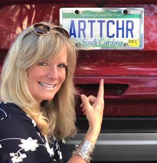A woman with blond hair and sunglasses on her head smiles while pointing to a license plate that says Arttchr on the back of a red vehicle.