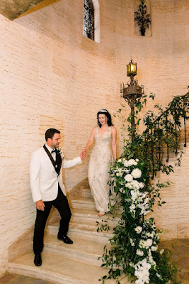 broom holding brides hand as she walks down staircase with handrail covered in flowers