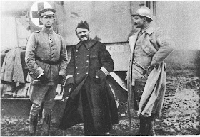 Black and white photo of three military commanders standing in front of a what appears to be a metal container with a cross sign on it. One on the very right is wearing a German military winter uniform and the one right beside in the center is wearing a French military trench coat. Another guy is standing on the very right side with a thick but light-colored winter coat with a hard hat.