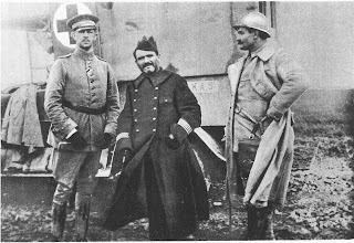 Black and white photo of three military commanders standing in front of a what appears to be a metal container with a cross sign on it. One on the very right is wearing a German military winter uniform and the one right beside in the center is wearing a French military trench coat. Another guy is standing on the very right side with a thick but light-colored winter coat with a hard hat.