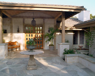 entrance porch area in a house that resembles her's: there's a wooden bench under a roofed area with columns by the door and the open area has a stone table and bench, some plants here and there