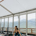 Woman in black top lifting barbell beside windows overlooking mountains