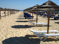 Na plaży na wschodnim krańcu wyspy Tavira. Piaszczysta rozległa plaża, na której zwartym rzędem stoją białe leżaki z towarzyszącymi im parasolami. W tle widać cieńką linię błękitrnego morza.