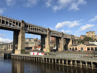 A view of the High Level Bridge showing the River Tyne in the foreground and a wooden structure with two huts on it by the feet of the bridge.  In the far right hand corner is an old stone building which is Newcastle Castle.  Photograph by Kevin Nosferatu for The Skulferatu Project.