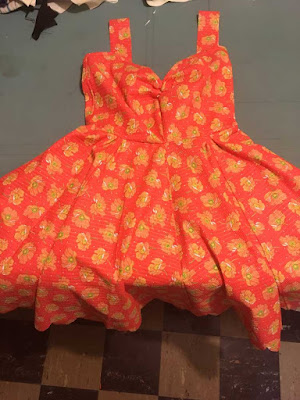 A bright coral sundress with a gathered sweetheart neckline and full skirt, and an orange poppy flower print, laid flat on a pale blue table with the hem of the skirt hanging over the edge.