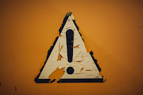 A torn and battered yellow triangular sticker with a black exclamation mark in the center is attached to an orange background, the corners of which are darkened.