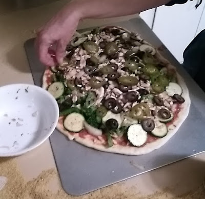 Pizza dough on pan, spread with sauce and toppings. In approximately 10-o'clock position on the roughly oval pizza, a hand adjusts a jalapeno.