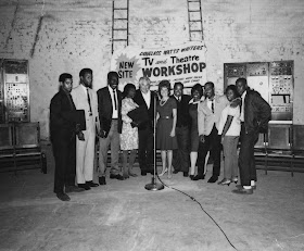 A photograph of a group of figures standing in front of a banner for a TV and Theatre workshop.