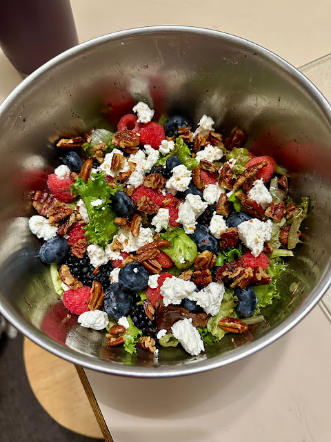 image of a stainless steel bowl filled with spring greens, goat cheese, fresh blueberries and raspberries, and spiced pecans