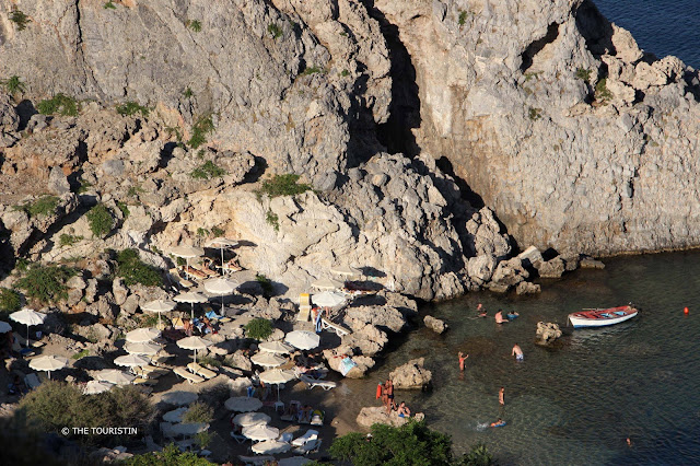 Tiny beach with sun umbrellas, a few people and a boat in the blue water, next to rocky cliffs..