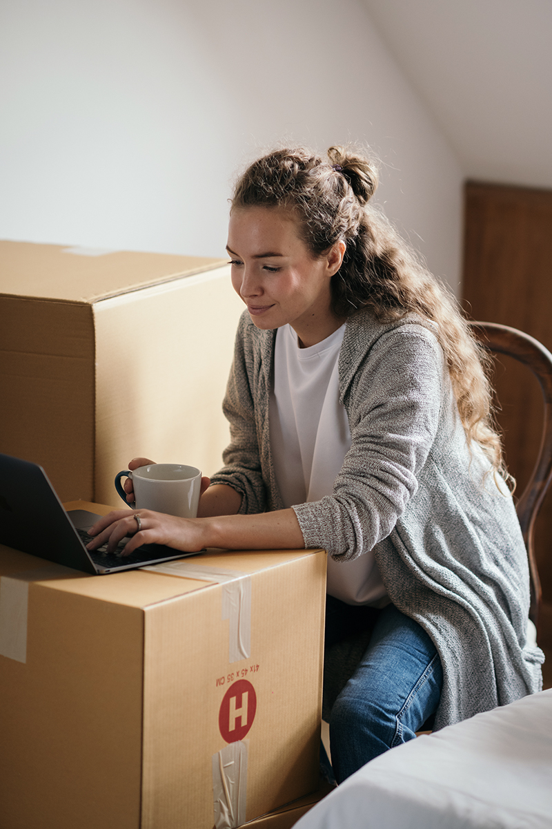woman with a laptop and coffee cup in her hand is sitting on a cardboard box