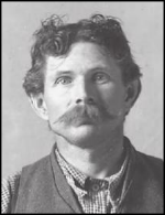 A white man in a plaid shirt and vest, looking head-on toward the camera. He has mussed short brown hair and a large mustache.