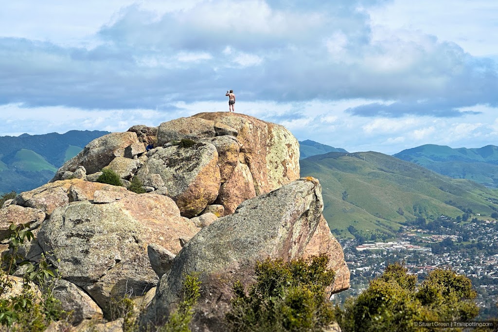 A man stands on the summit, dwarfed by the massive bare rocks of Bishop Peak Summit.