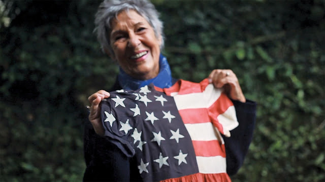 An older woman with short hair holds up a dress her made her as a child.