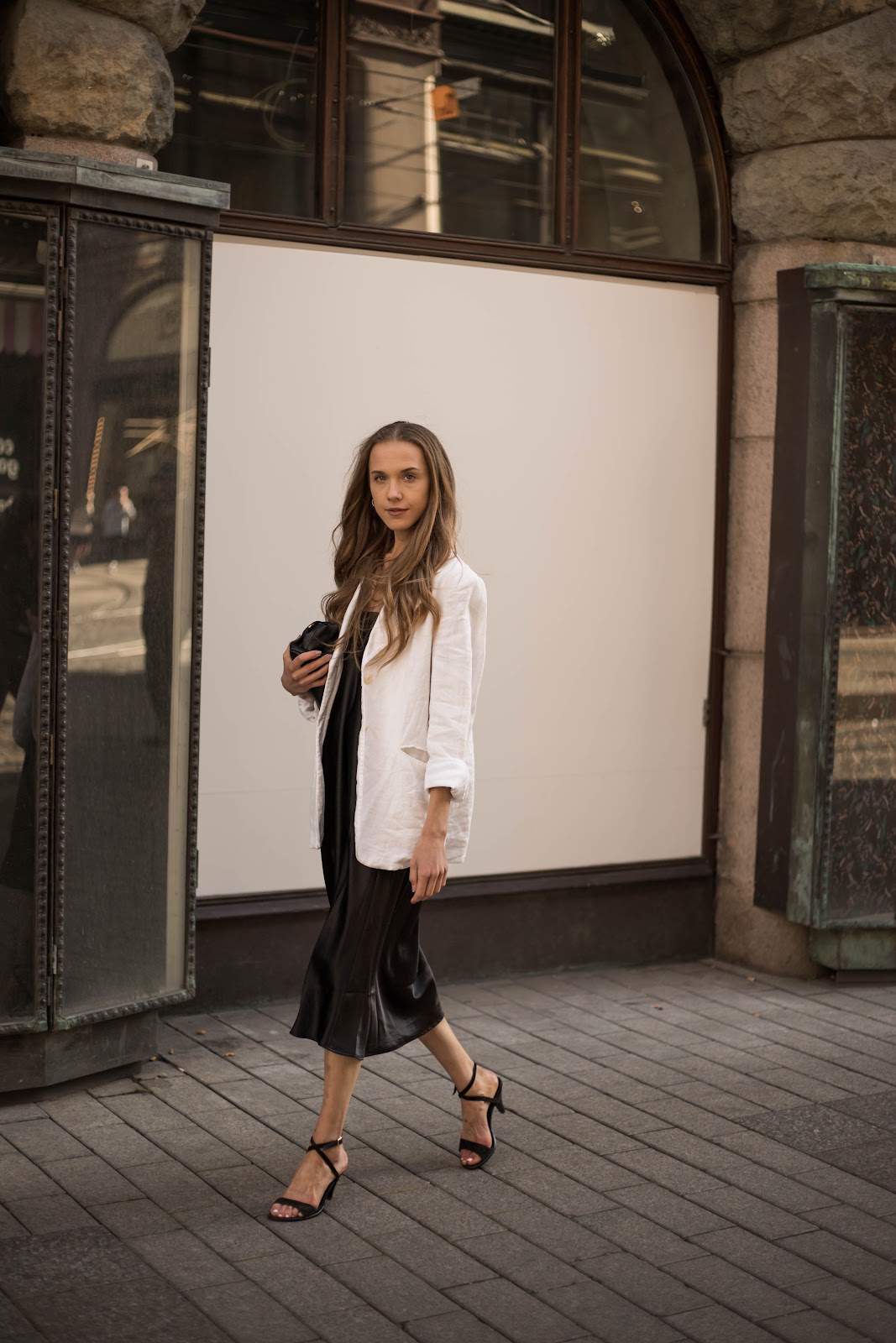 Nainen kävelee kadulla päällään musta satiinimekko, valkoinen bleiseri, mustat korkokengät ja musta pikkulaukku