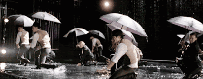 the glee kids performing on a flooded stage with umbrellas