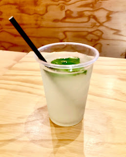 A cylindrical clear cup filled with cloudy lenonade with a piece of bright green oval mint with a red and white cylindrical straw on a light brown rectangular table on a bright background