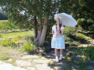 Me wearing a causal lolita coordinate with blues, pinks and whites. My skirt is blue. It is an oldschool coordinate. I am standing in the shade and holding a parasol in a sunny garden.