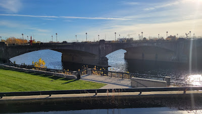 Photo looking across a grassy lawn at a river. A bridge with multiple low arches carries vehicular traffic over the river.