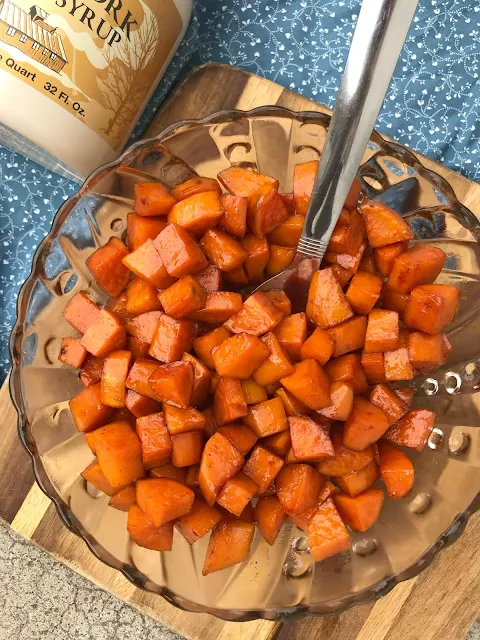 Serving bowl of glazed sweet potatoes and a bottle of maple syrup.