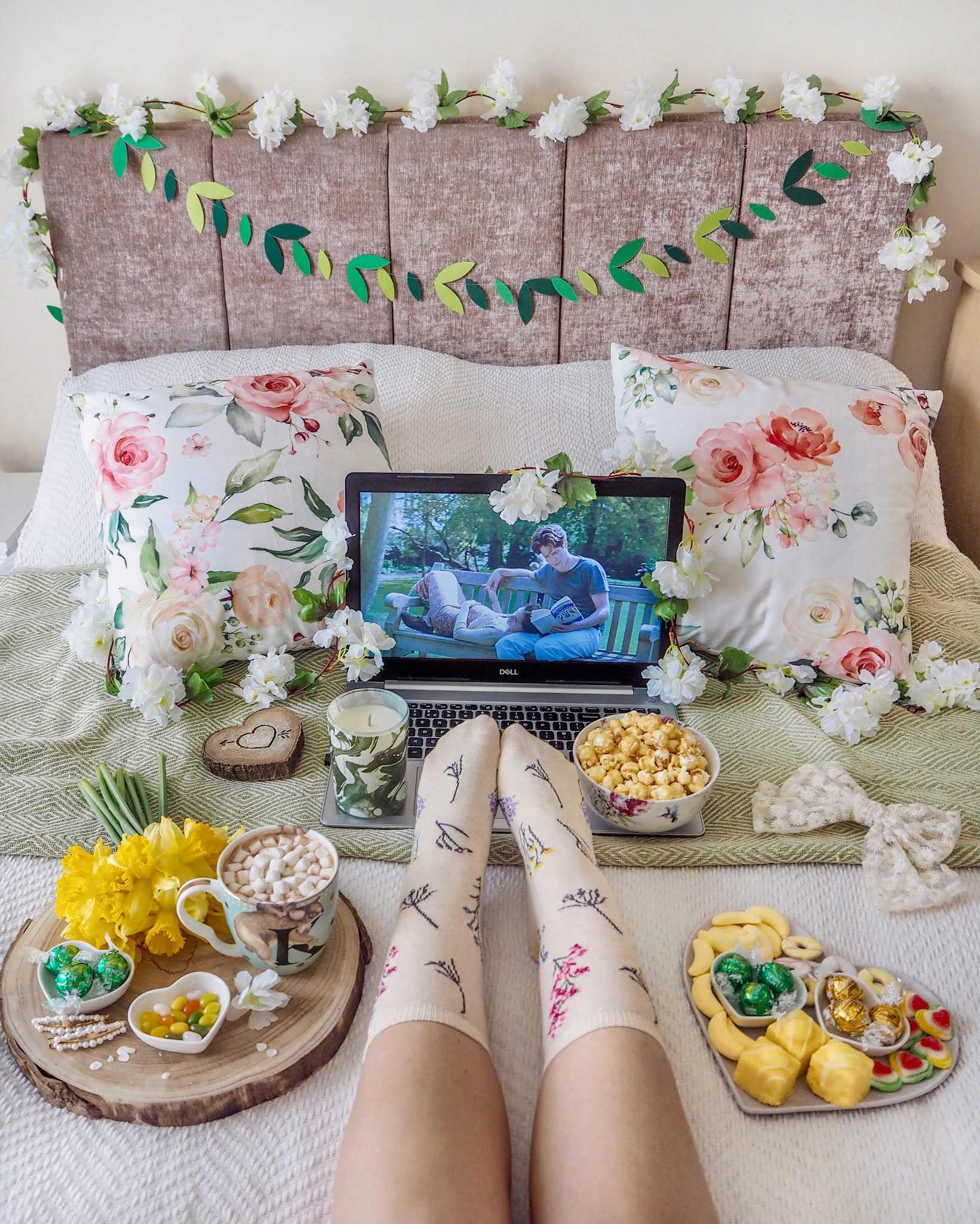 A cosy movie night shot watching Notting Hill on a laptop on a bed with lots of green and yellow floral spring props surrounding. You can see the girls legs on the bed wearing floral socks