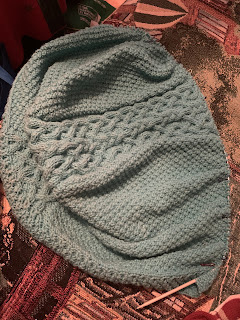 The hood laid out somewhat flat. The shaping makes it drape over a bit in the center. The star stitch border, center cable panel, and moss stitch of the sides are all very clear, both in terms of their textures and differences to one another.