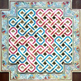 The board at the end of a game. The board is square, with the corners cut out so it forms a sort of cross shape. Along the edge of the board is complex celtic knotwork in an elaborate zoomorphic theme with birds entwined. The play area itself is filled with plastic pieces; these are rings in both blue and red which are joined to the pieces next to them with 'bridges' that combine the rings into a celtic knotwork pattern.