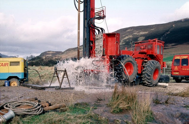 Bridge Farm borehole, Machrie, Arran. Drilling a groundwater production borehole for West of Scotland Water at Machrie, Arran.  The drill rig is located close to the 'String' road from Brodick at a site chosen by the Hydrogeology Group of BGS. The rig is using an air flush hammer to drill through Permian sandstone. The compressor is seen at the left. Compressed air is sent down the drill rods to the hammer at the bottom of the borehole. The air operates the hammer and brings cuttings and water to the surface. The water flows away from the site to a stream. The 4-wheel drive tractor unit powers the rig and is highly manoeuvrable - useful for sites with problematical access.